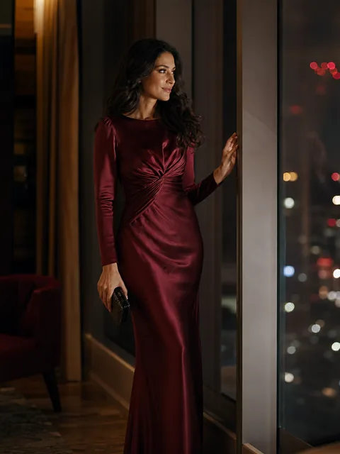 An adult woman in a burgundy satin evening gown standing by a tall window of a hotel lounge at night, city lights behind her