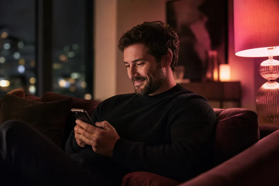 An adult man relaxing on a sofa at night smiling at his phone, a warm lamp and a city window behind him