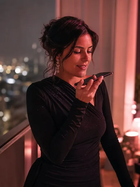 An adult woman in an elegant dress recording a voice message on her phone on a night balcony